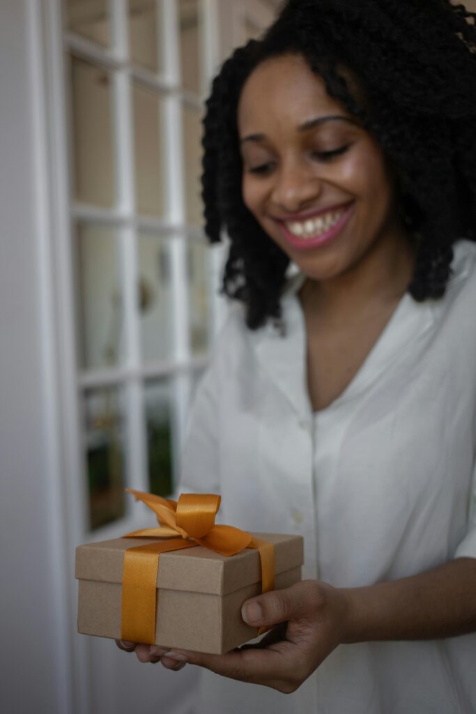 An African American woman joyfully holds a gift box wrapped with an orange ribbon indoors.